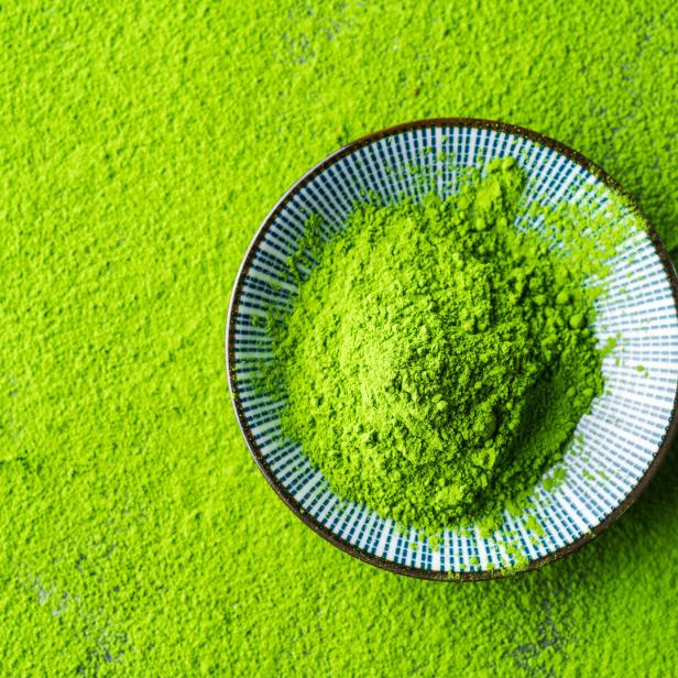 Eine kleine Schale mit leuchtend grünem Matcha-Pulver steht auf einer Fläche, die ebenfalls mit Pulver bedeckt ist.