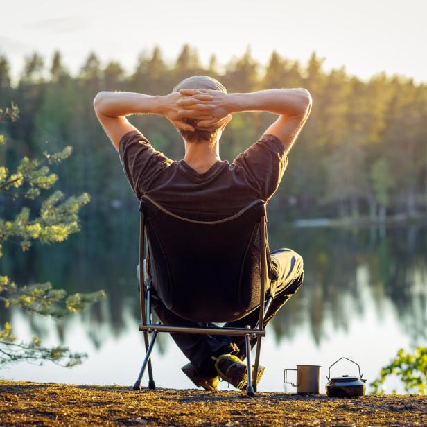 Man is sitting in a camping chair on the background of a forest lake on a beautiful summer evening.