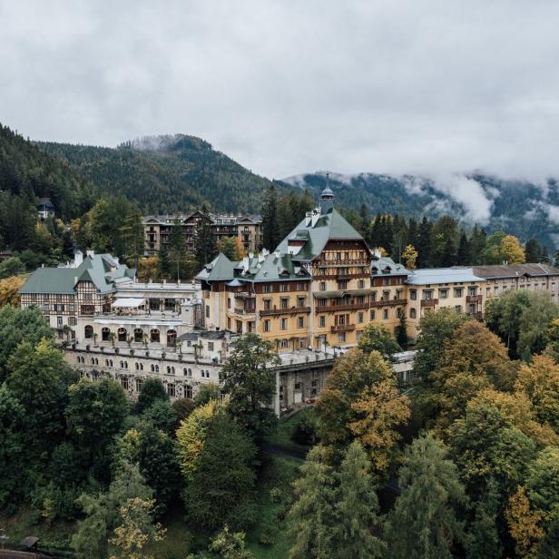 Das große Südbahnhotel mit seinen verschiedenen Trakten und großem Balkon. Es thront auf einem bewaldeten Hang. Im Hintergrund erheben sich Berge, Nebel lichtet sich