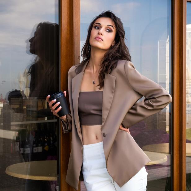 Ein attraktives Model posiert vor einem Café mit einer Tasse Kaffee in der Hand und trägt ein Bandeau-Top, eine weiße Hose und ein Sakko.