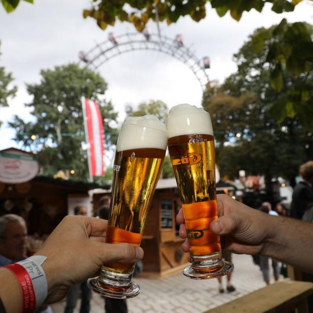 Zwei Hände mit Biergläsern, die gerade zusammenstoßen, im Hintergrund sieht man das Wiener Riesenrad