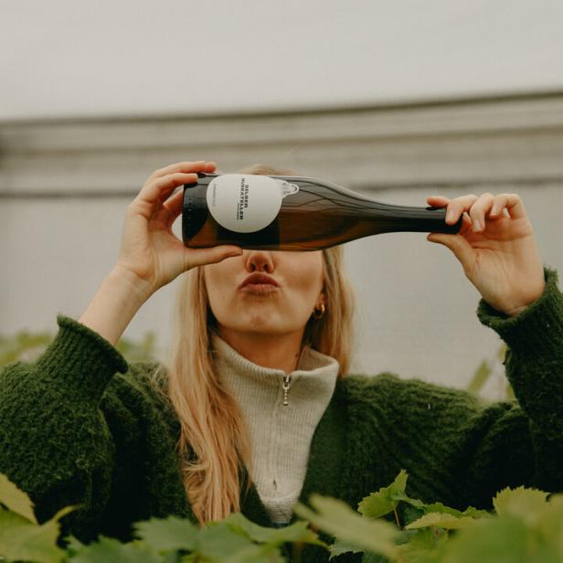 Winzerin Katharina Gessl steht zwischen ihren Weinstöcken, hält eine ihrer Weinflaschen vors Gesicht und zeigt das Etikett. 