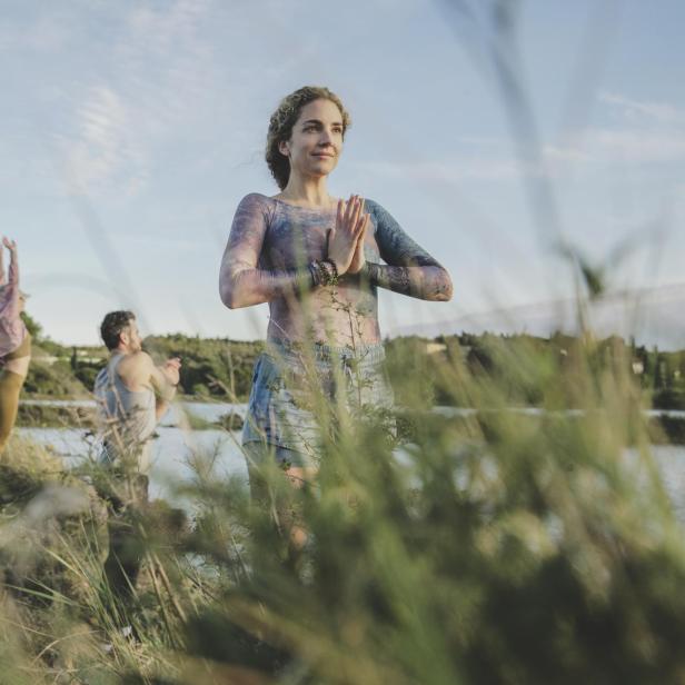 Drei Personen machen Yoga im Grünen, im Hintergrund das Meer.