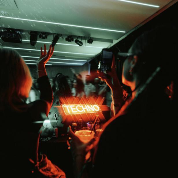 Blick vom DJ-Pult auf den Innenraum. Menschen haben die Hände in der Höhe und tanzen, eine Person fotografiert.. Auf einem Leuchtschild steht Techno.