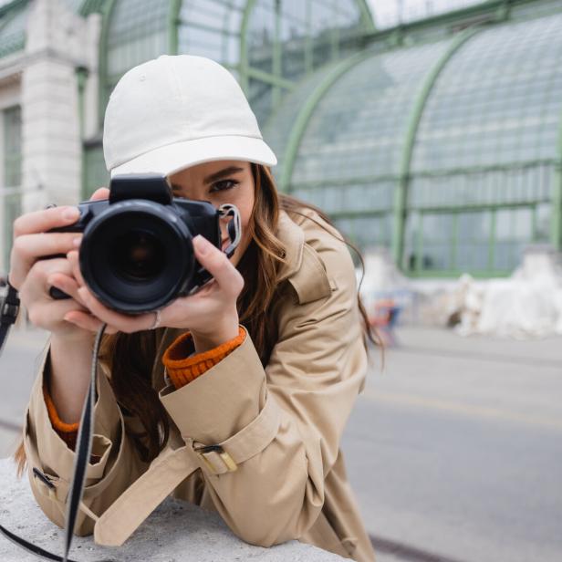 Eine Frau mit beiger Jacke und weißer Kappe hält eine Kamera und fotografiert vor dem Palmenhaus im Burggarten in Wien.