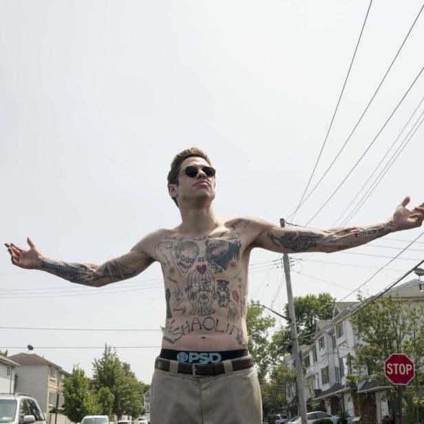 Szene aus dem Film The King of Staten Island (2020): Pete Davidson steht mit nacktem Oberkörper, der mit Tattoos bedeckt ist, und ausgebreiteten Armen und Sonnenbrille auf einer Straße in einem Wohngebiet.