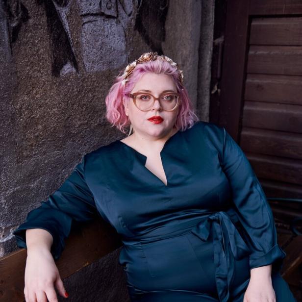 46-219400461 Bobby Herrmann-Thurner sitzt auf einer Bank, trägt einen dunkelblauen Jumpsuit und rosa Haare.