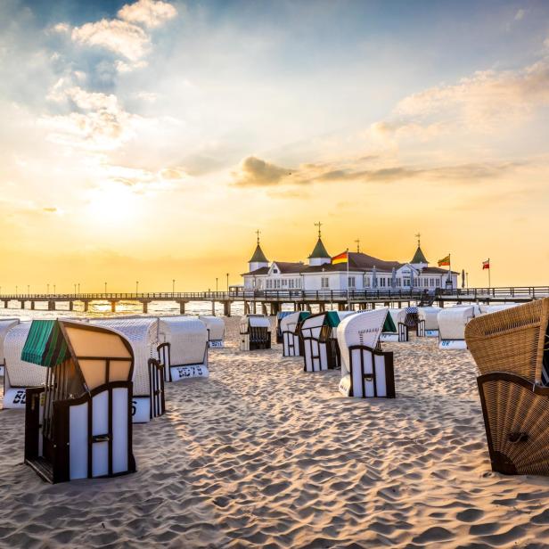 Strand mit mehreren Strandkörben in verschiedenen Farben und einer Seebrücke mit Gebäude im Hintergrund bei Sonnenuntergang.