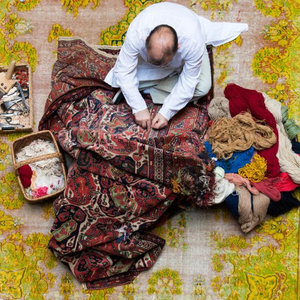 Vogelperspektive: Ein Mann hockt am Boden, vor ihm ein Teppich, neben ihm Wolle und Flickzeug. Er arbeitet am Perser mit klassischem Muster.