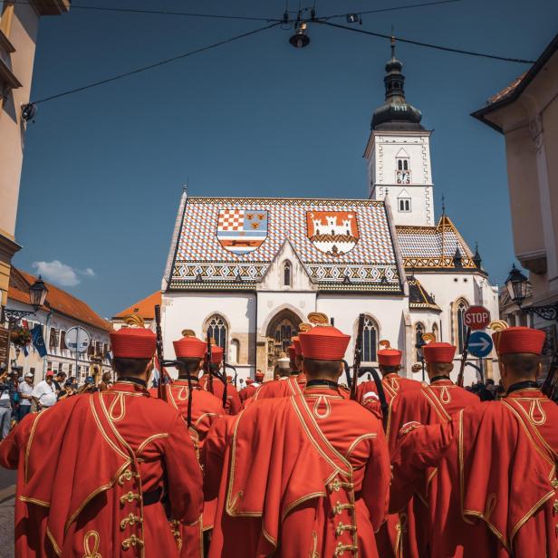 Gruppe von Geistlichen in roten Uniformen mit goldenen Verzierungen vor der St.-Markus-Kirche in Zagreb mit farbigem Dach und Turm.