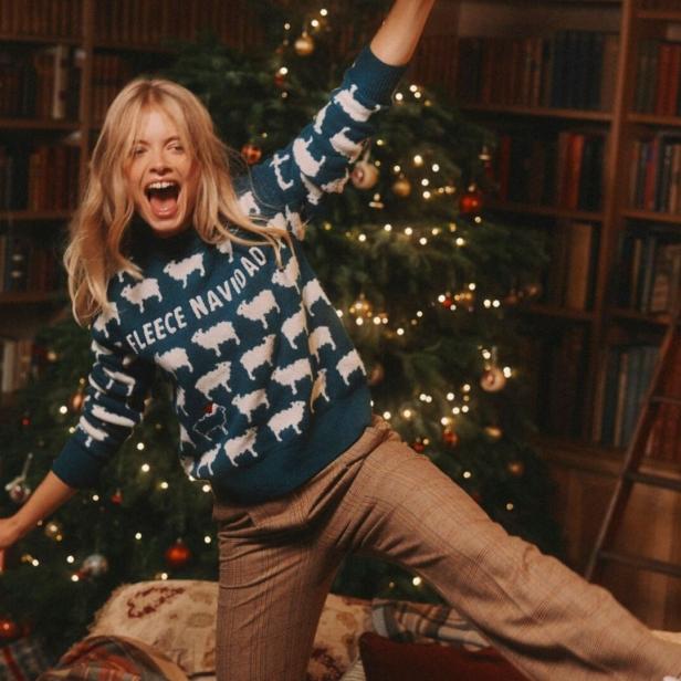 Eine blonde Frau im Weihnachtspullover mit Schafen jubelt vor einem Weihnachtsbaum und Bücherregalen.