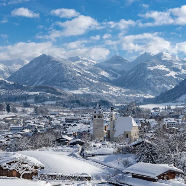 Blick von oben ins Tal auf Kitzbühel: Zwei Kirchen ragen heraus, dahinten erheben sich die Berge. Die Landschaft ist verschneit.