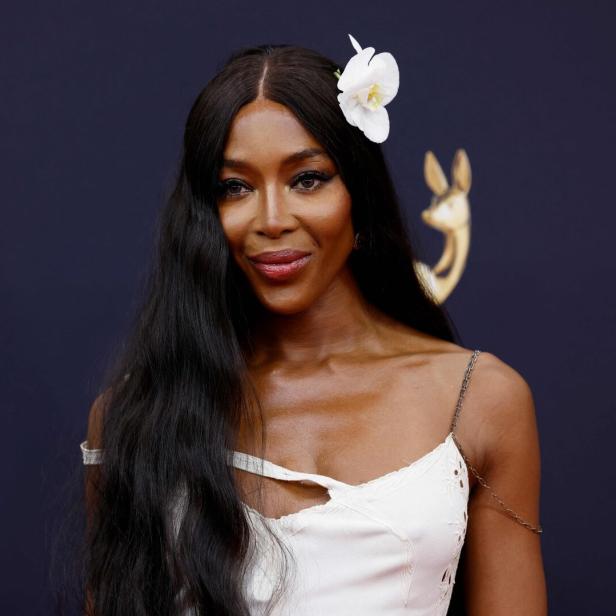 Naomi Campbell mit weißer Blüte im Haar und weißem Kleid vor dunklem Hintergrund.