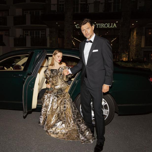 Ein Mann und eine Frau, die aus einem Auto steigen. Er trägt einen Smoking, sie ein goldenes Kleid. 