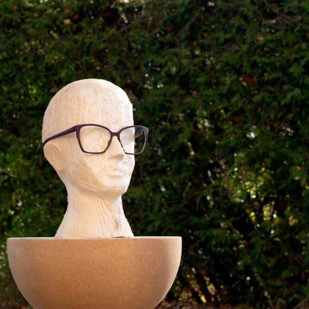 Holzskulptur eines Kopfes mit Brille, stehend in einer großen, runden Schale vor grünem Laub.