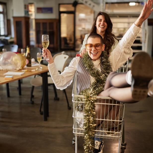 Zwei lachende Personen feiern ausgelassen, eine sitzt mit Sektglas und Girlande in einem Einkaufswagen, die andere schiebt sie.
