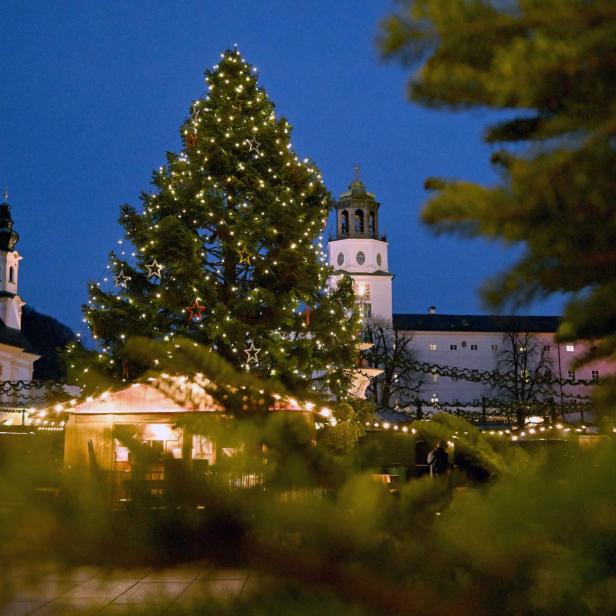 Ein Weihnachtsbaum mit Lichtern und Sternen steht auf einem Platz in Salzburg vor Gebäuden.
