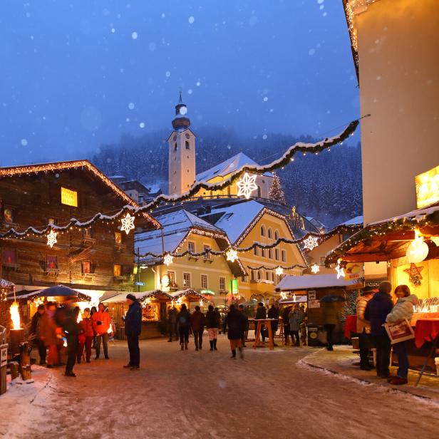 Weihnachtsmarkt mit beleuchteten Holzständen, schneebedecktem Boden und Kirche im Hintergrund bei Abenddämmerung.