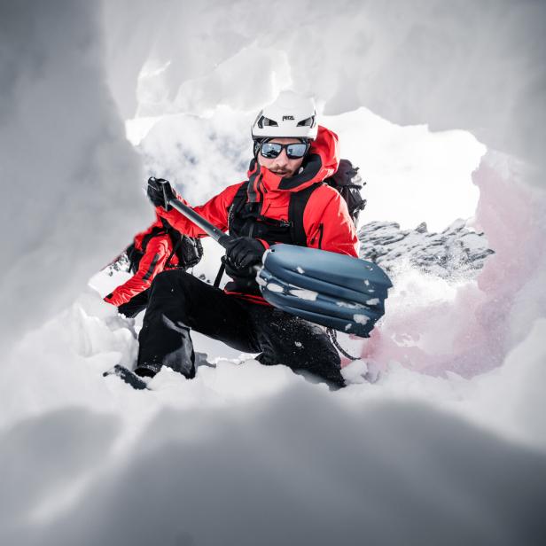 Zwei Personen in roter Winterkleidung mit Skiausrüstung in einer Schneehöhle, umgeben von Eiswänden.