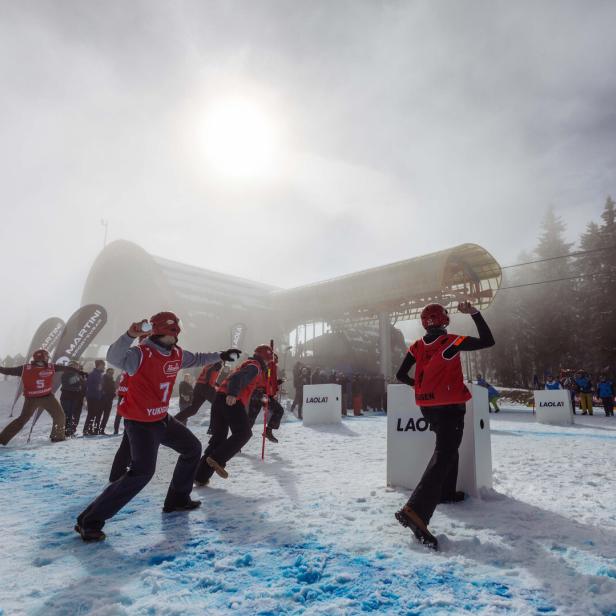 Mehrere Personen in roter Skibekleidung und Helmen stehen auf schneebedecktem Boden vor einer großen, weißen Schneekanone unter bewölktem Himmel.