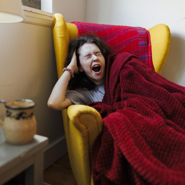 Eine Frau gähnt müde in einem gelben Sessel unter einer roten Decke.