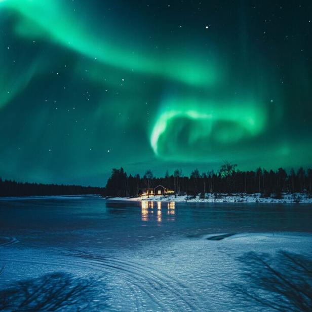 Grünes Polarlicht leuchtet am Nachthimmel über einem zugefrorenen See mit beleuchtetem Haus am Ufer.
