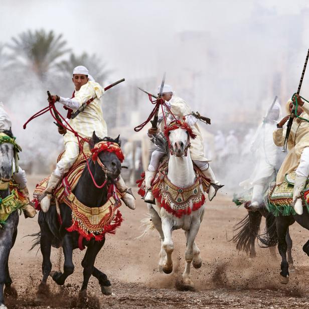 Mehrere Reiter in traditioneller Kleidung reiten auf geschmückten Pferden und halten Gewehre in die Luft, Staub wirbelt auf.