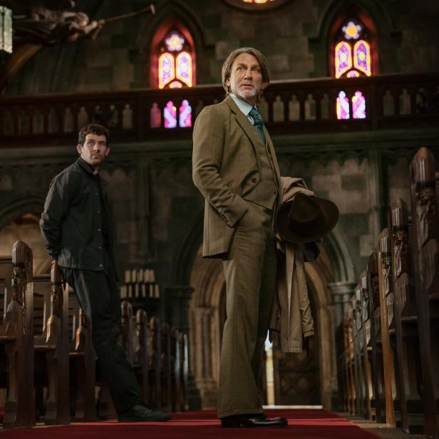 Zwei Männer stehen zwischen Holzbänken in einer Kirche mit bunten Fenstern und schauen in verschiedene Richtungen.