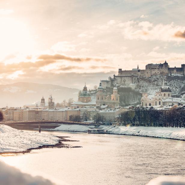 Verschneite Stadt mit Fluss, historischen Gebäuden und einer Burg auf einem Hügel im Sonnenlicht.