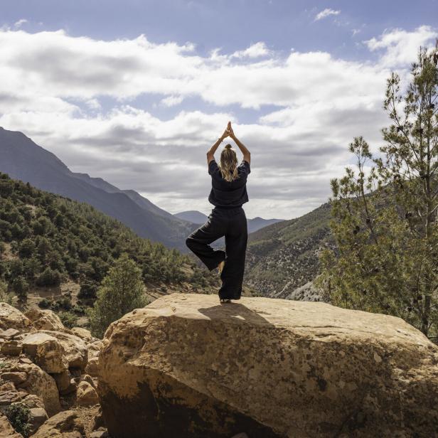 One woman practising yoga in tree pose on rock overlooking forested mountains in nature in the Atlas Mountains, Ourika Valley, Morocco