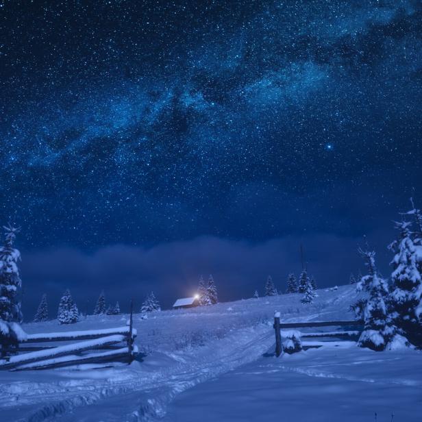 Verschneite Landschaft mit Tannen, einer Hütte mit Licht und einem klaren Sternenhimmel in der Nacht.