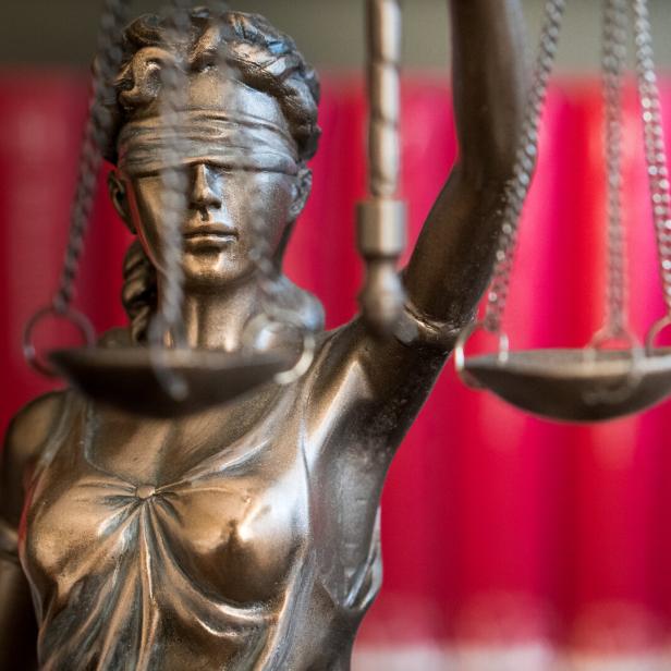 Eine Justitia-Statue mit Augenbinde und Waage steht vor einem roten Hintergrund.