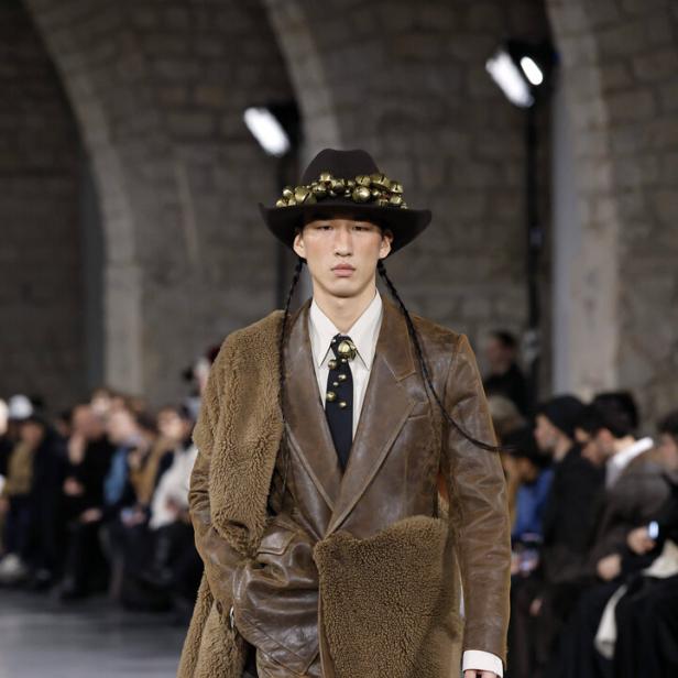 Ein Model mit Hut und Zöpfen präsentiert braune Lederkleidung auf einem Laufsteg.