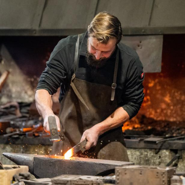 Ein Schmied mit Bart bearbeitet glühendes Metall auf einem Amboss mit Hammer.