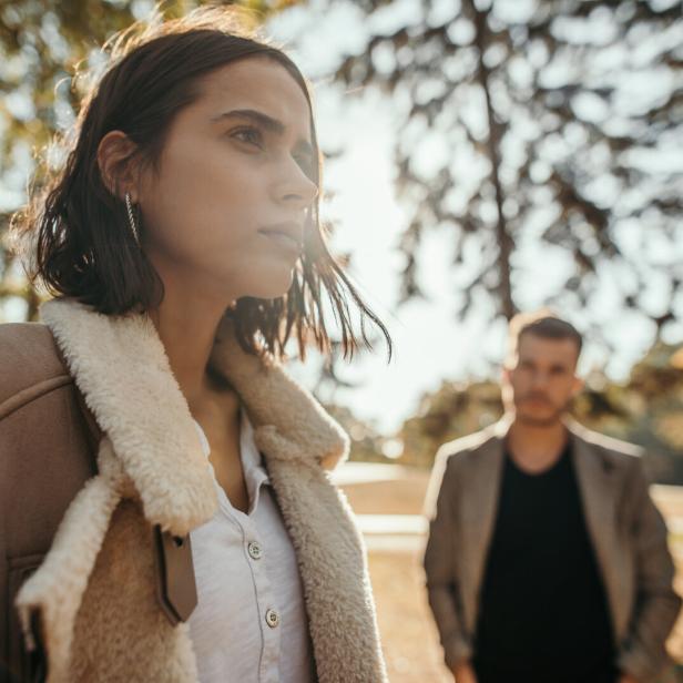 Eine Frau mit brauner Jacke und Fellkragen schaut besorgt aus, ein Mann steht im Hintergrund.
