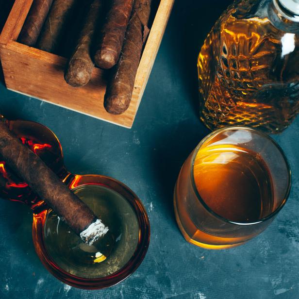 Eine brennende Zigarre liegt in einem Aschenbecher neben einem Glas und einer Karaffe mit braunem Getränk sowie einer Zigarrenkiste.