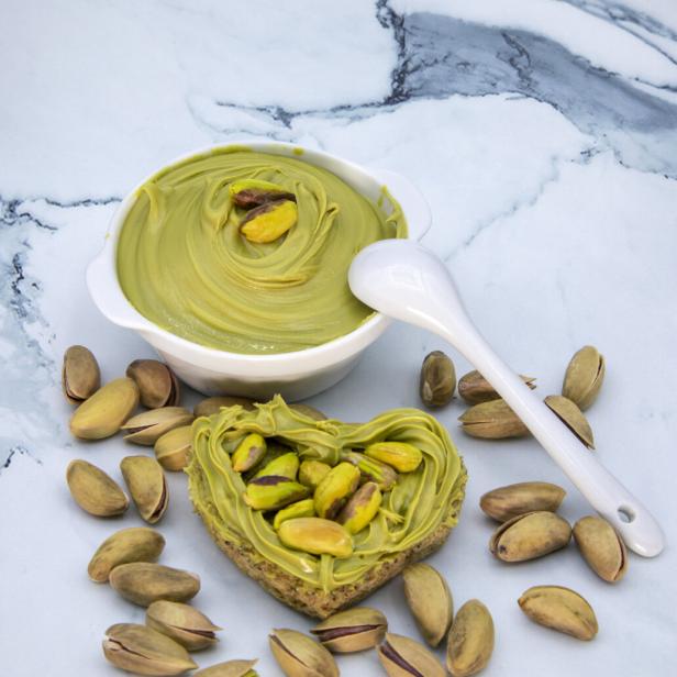 Herzförmiges Brotstück mit Pistaziencreme und Pistazien, daneben eine Schale mit Creme und ein Löffel, umgeben von Pistazien.
