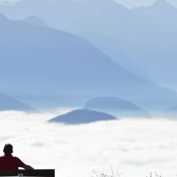 Eine Person sitzt auf einer Bank und blickt auf eine weite Berglandschaft im Nebel.