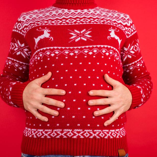 Eine Person im roten Weihnachtspullover hält sich den Bauch vor rotem Hintergrund.