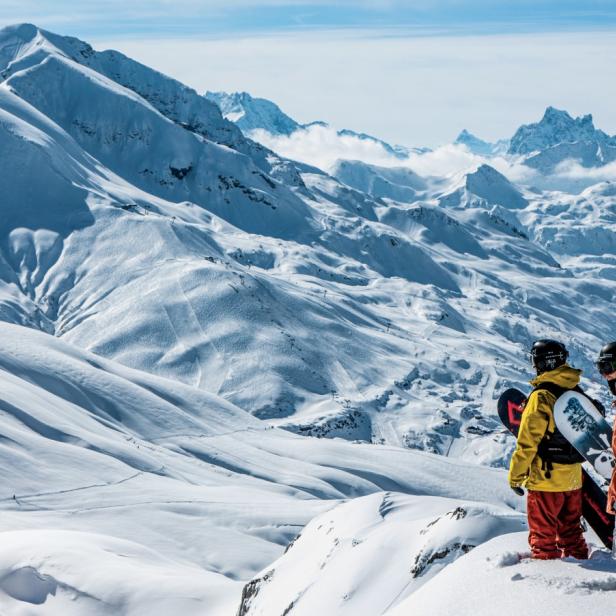 Zwei Snowboarder stehen auf einem verschneiten Gipfel im Skigebiet Arlberg und blicken auf die imposanten Alpen mit langen, leeren Abfahrten.