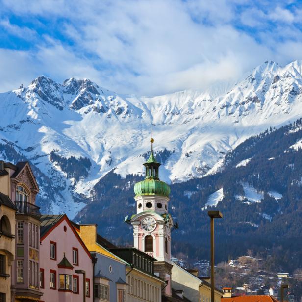  Bunte Hausfassaden und ein Kirchturm heben sich vor einer winterlichen Bergkulisse ab