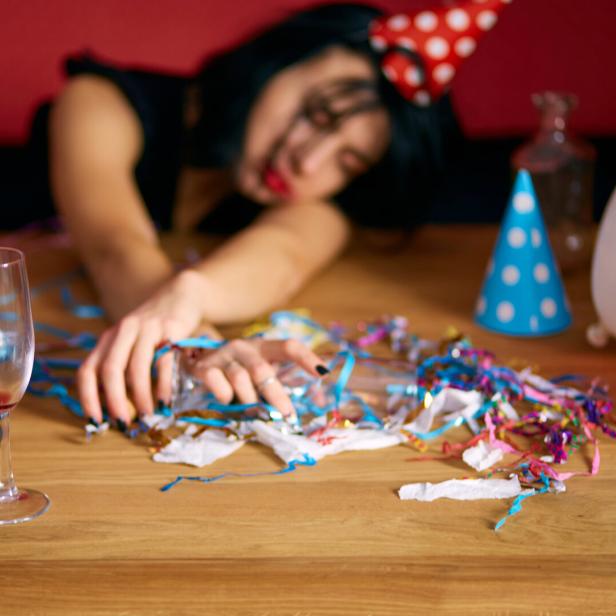 Eine Frau mit Partyhut schläft auf einem Tisch voller Konfetti und Luftschlangen, vor sich ein leeres Sektglas.