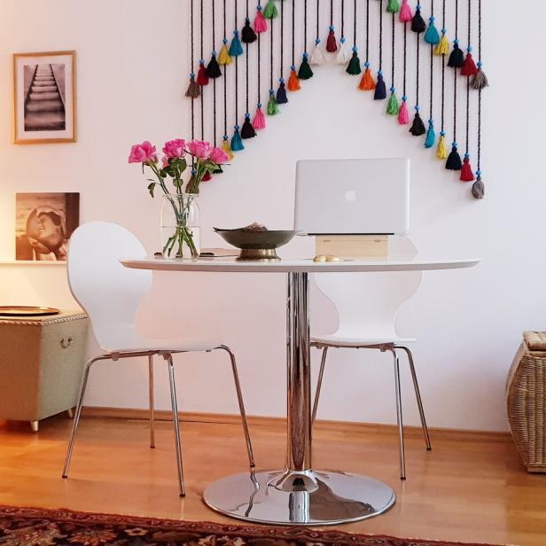 Runder Tisch mit zwei weißen Stühlen, darauf ein Laptop und Blumen, daneben bunte Wanddeko und Bücherregal.