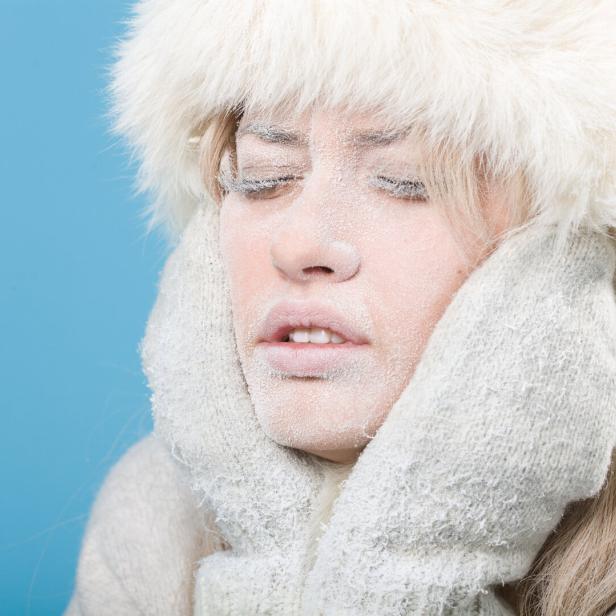 Eine Frau mit frostbedecktem Gesicht trägt eine Pelzmütze und Handschuhe vor blauem Hintergrund.