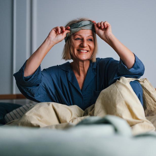 Eine lächelnde Frau im blauen Pyjama sitzt im Bett und zieht sich eine Schlafmaske vom Kopf.