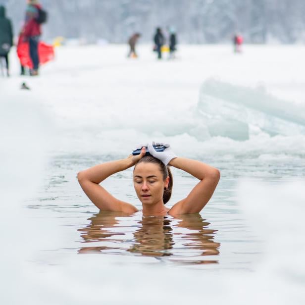 Eine Frau badet in einem eisigen See, umgeben von Schnee, und hält ihre Hände mit Handschuhen auf dem Kopf.