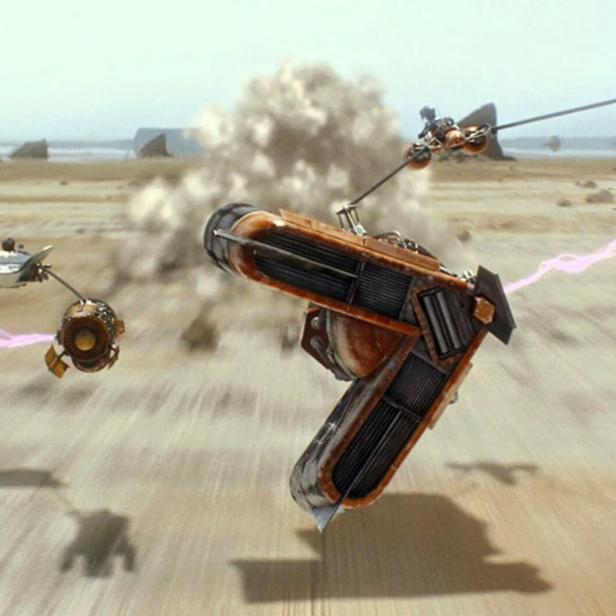 Zwei futuristische Rennfahrzeuge rasen mit hoher Geschwindigkeit über eine sandige Wüstenlandschaft.