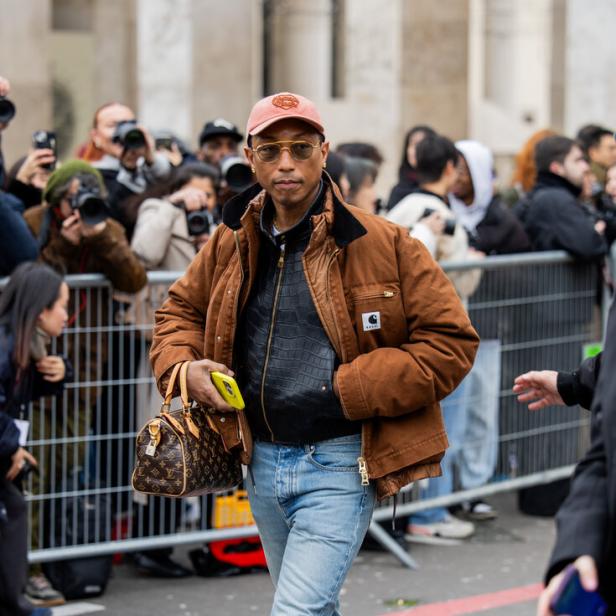 Pharrell Williams trägt eine braune Jacke, Schlaghosen und eine Louis Vuitton Tasche.