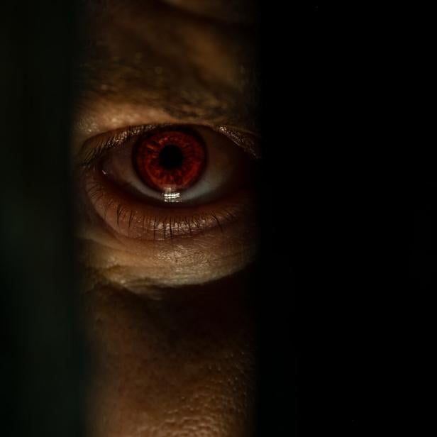 Ein rotes menschliches Auge blickt durch einen schmalen dunklen Spalt.