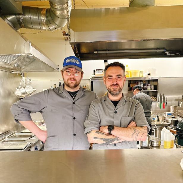 Zwei Köche in grauen Jacken stehen nebeneinander in einer professionellen Restaurantküche.